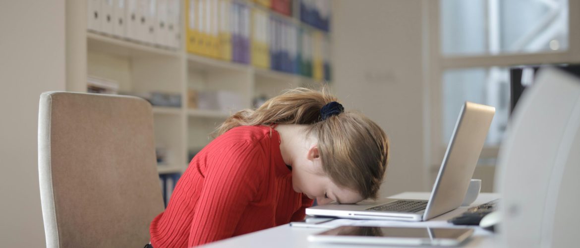 A tired woman in a red sweater leans her head on a desk with a laptop, symbolizing workplace fatigue.