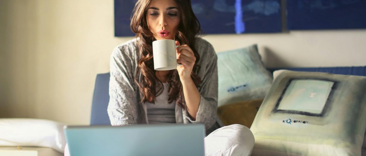 A woman enjoying coffee while working from home in a cozy bedroom setting with a laptop.